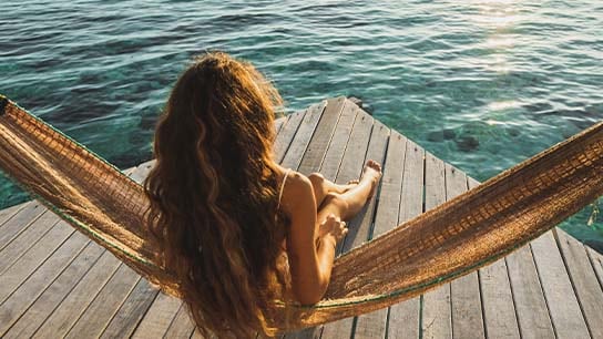 A woman appears sitting in a hammock while looking at the sea